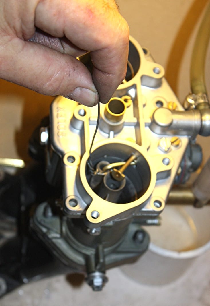 A hand holding a thin wire gauge against the throttle barrel of a Solex carburetor on a workbench, with the flange, butterfly shaft, and Solex casting visible.