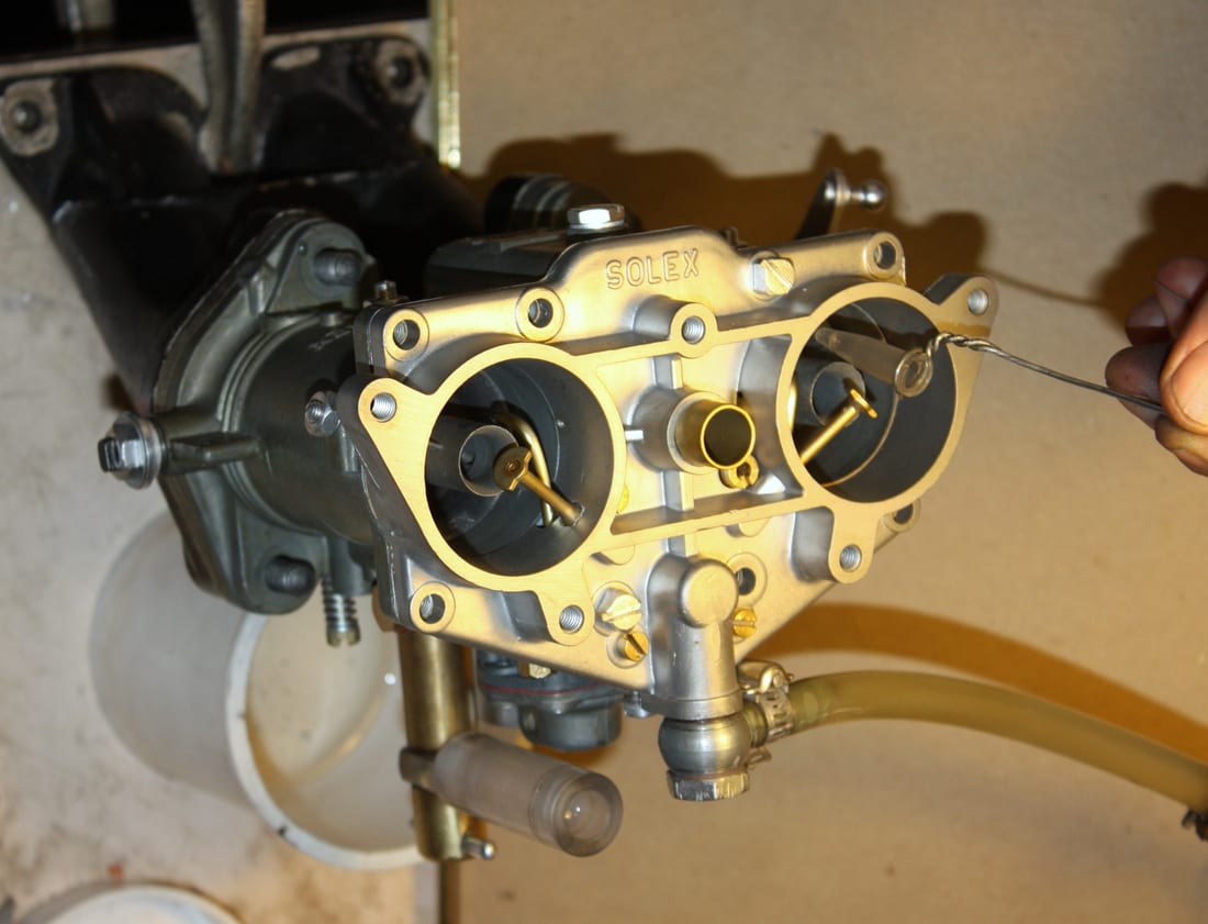 A hand inserting a thin wire gauge into the top of a twin-barrel Solex carburetor on a stand, showing both throttle bores and the Solex casting.