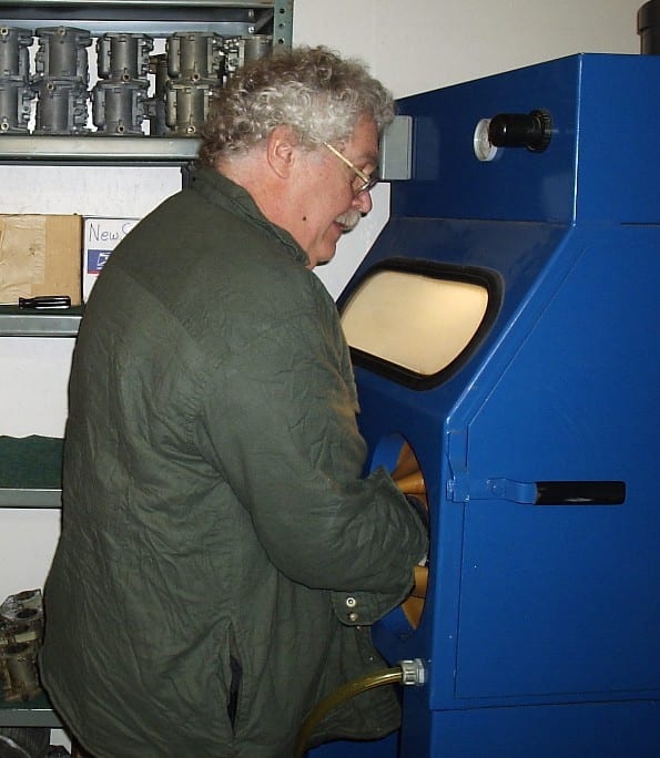 A man working at a bead-blasting cabinet with a rack of Zenith 32 NDIX carburetors behind him.