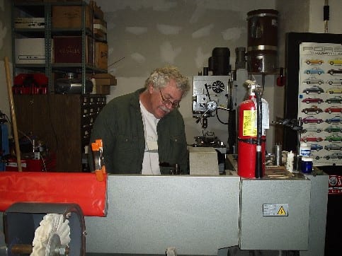 A man working at a restoration bench with a Porsche poster on the wall.